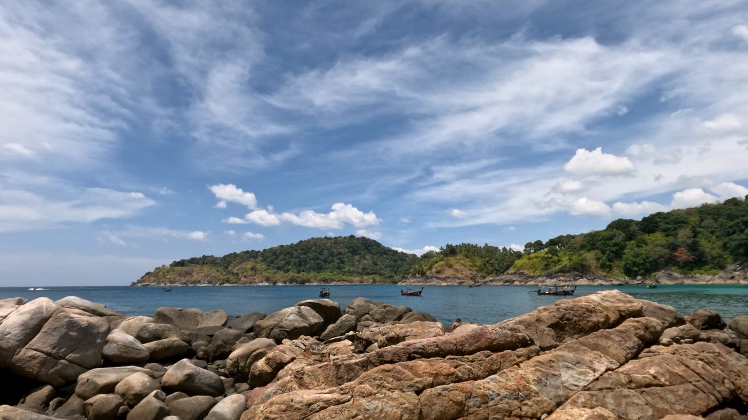 Longtail boats anchored off a rocky headland on the Phuket coast under a wide blue sky