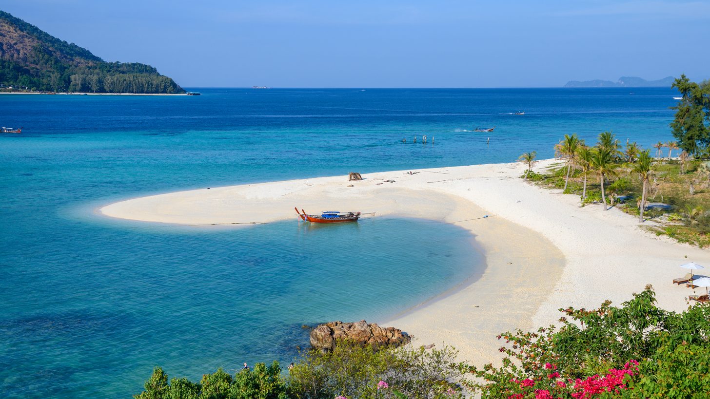 A longtail boat moored at a white sandspit on Koh Lipe with palms and clear shallow water