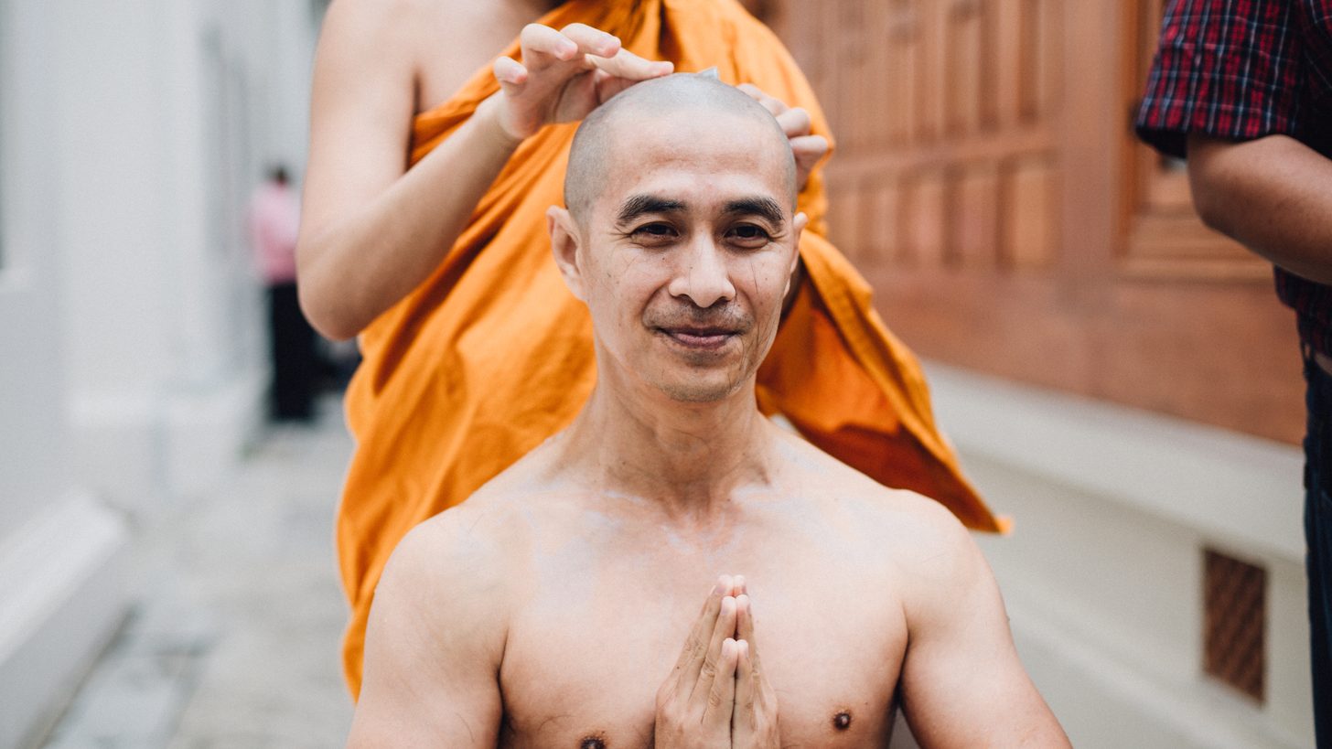 A man's head being shaved by a monk in orange robes at a Thai ordination ceremony