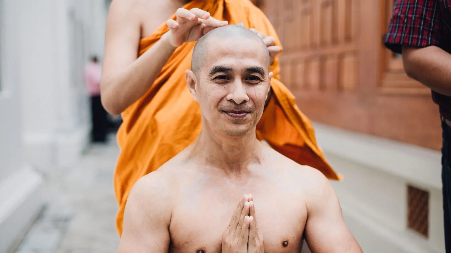 A man's head being shaved by a monk in orange robes at a Thai ordination ceremony