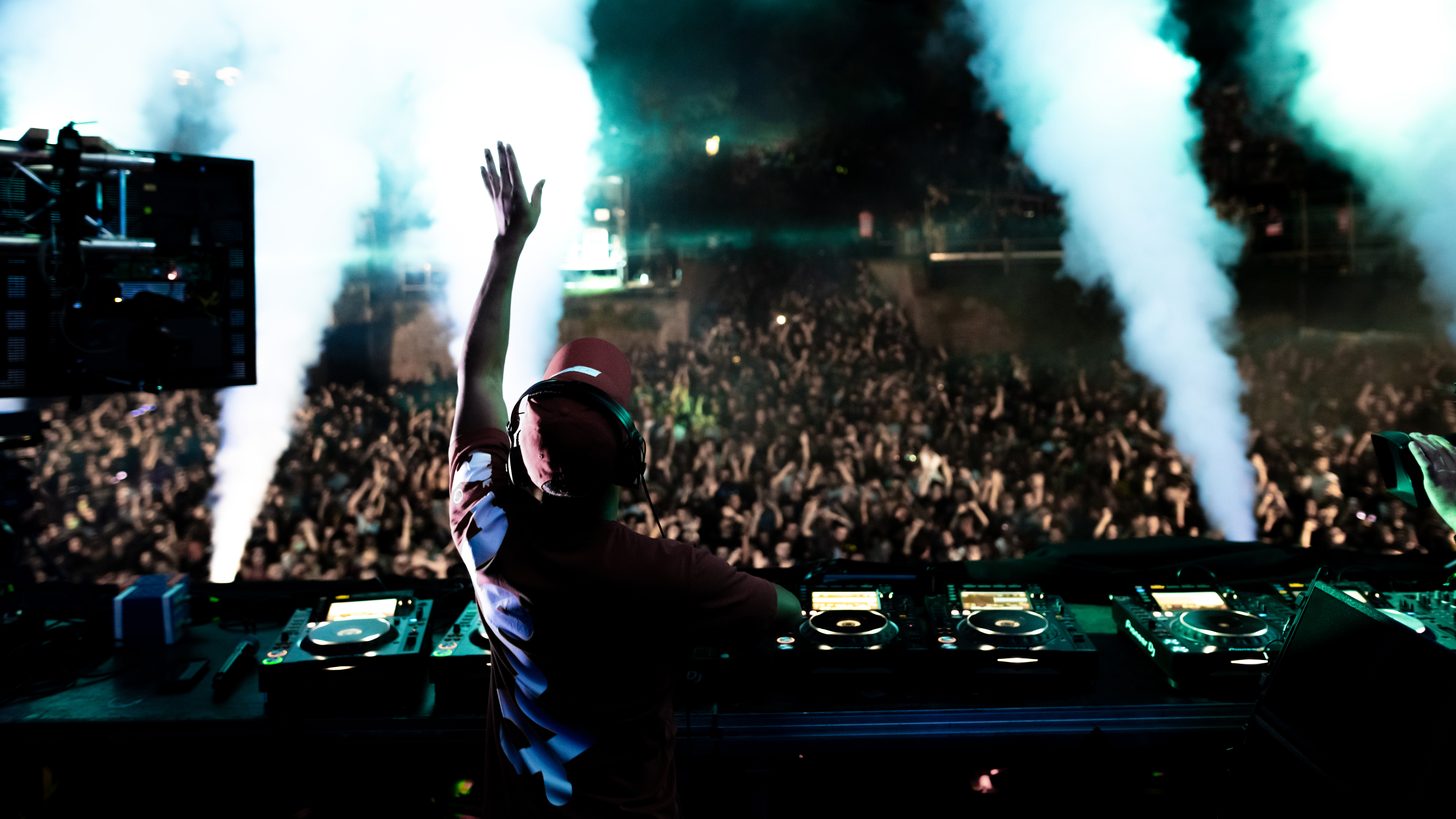 A DJ performing to a large illuminated festival crowd at night