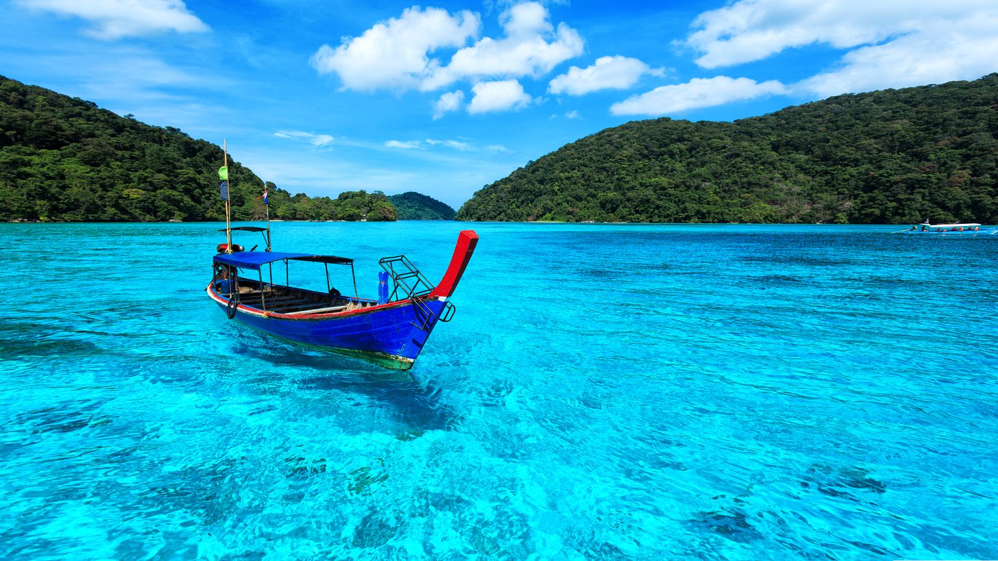Ein Longtail-Boot fährt bei teils bewölktem Himmel über ruhiges türkisfarbenes Wasser nahe einer Insel im Golf von Thailand