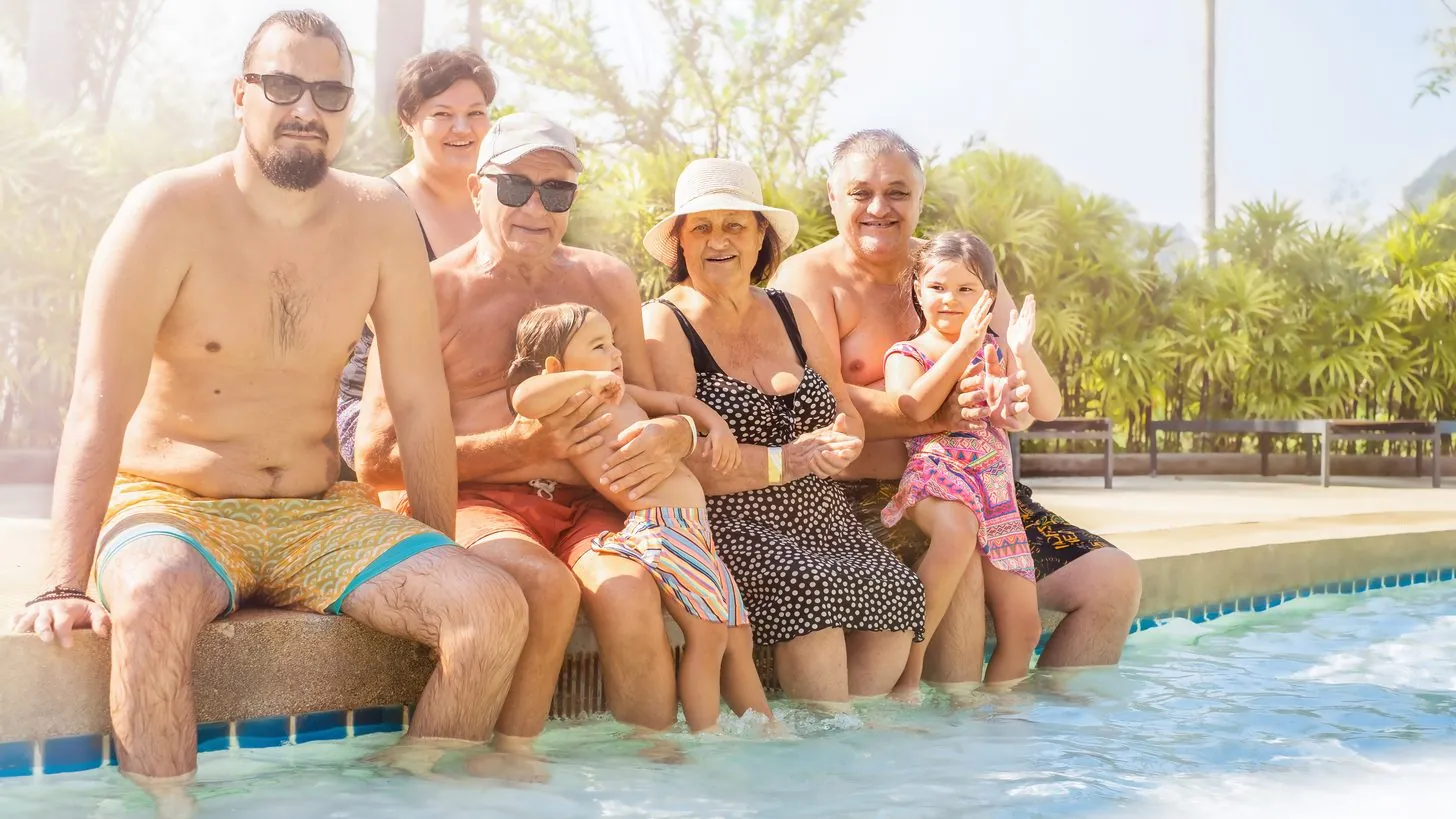 Drei Generationen einer Familie beim gemeinsamen Frühstück am Pool auf der Terrasse einer privaten Villa in Phuket — die Großeltern sitzen im Schatten, die Kinder dahinter am Beckenrand