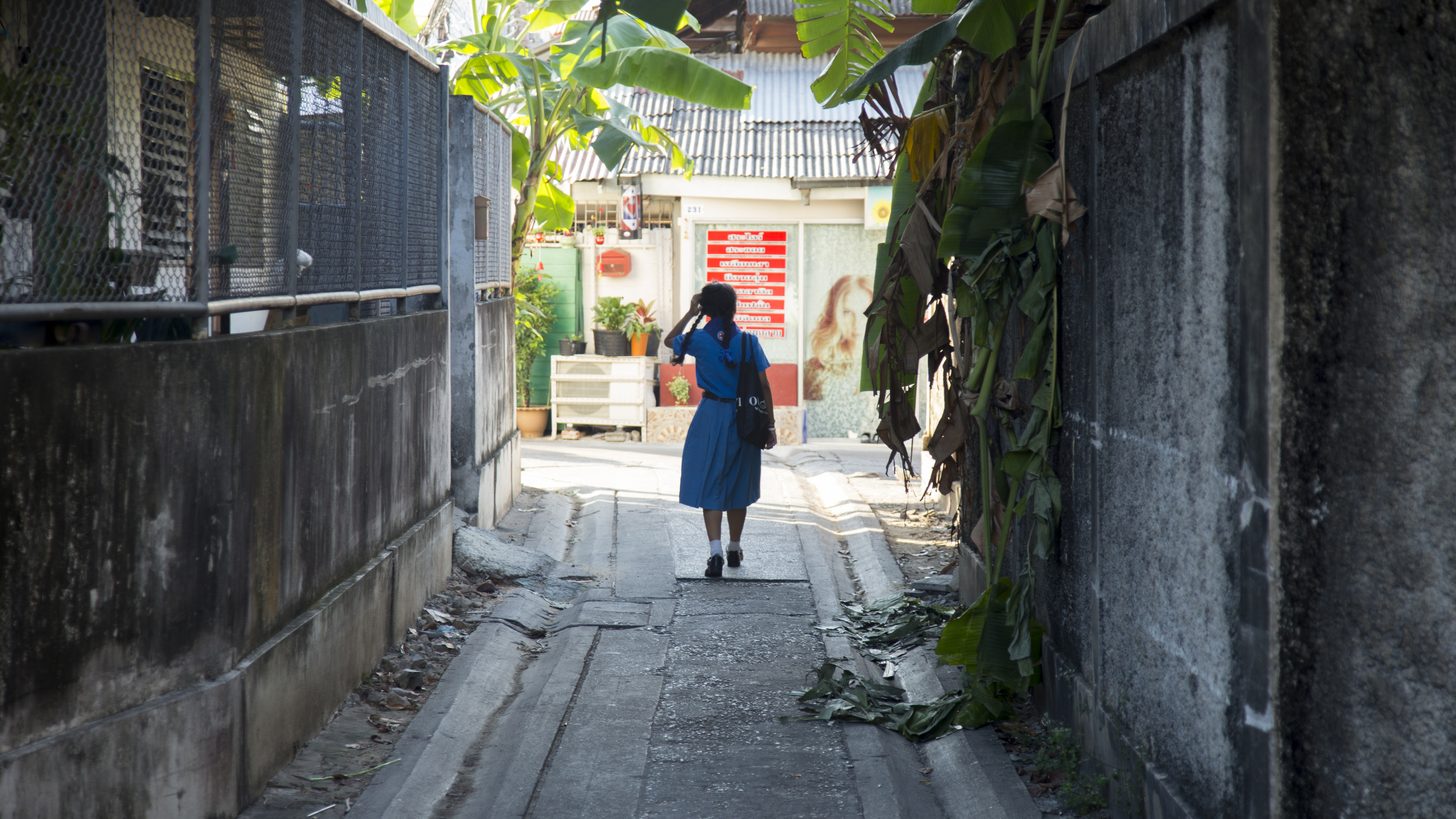 Une ruelle étroite entre murs et clôtures dans un quartier riverain de Bangkok, large seulement pour les piétons, une silhouette marchant vers les commerces du fond