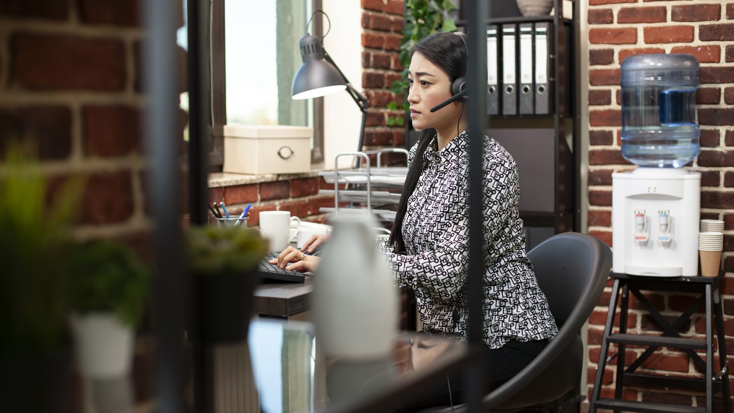 A contact centre operator wearing a headset at a desk, taking a call in front of a computer screen