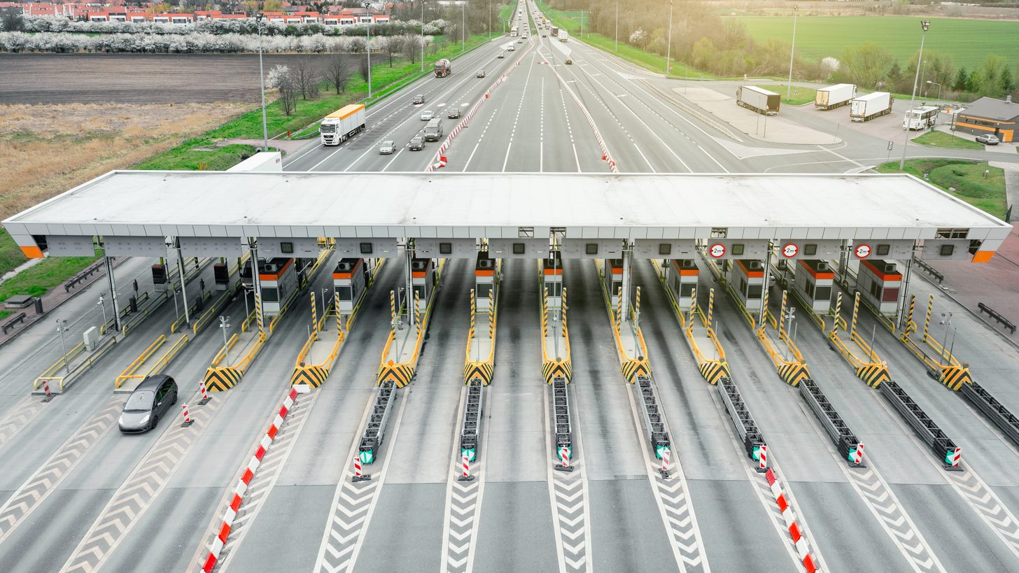 Vehicles queuing at a manned road checkpoint booth beneath a covered canopy