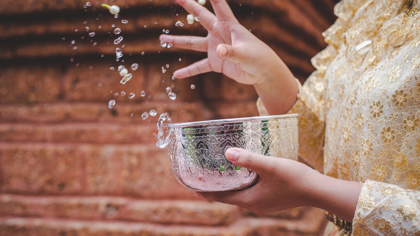 A hand pouring water from a bowl during a Songkran blessing at a temple