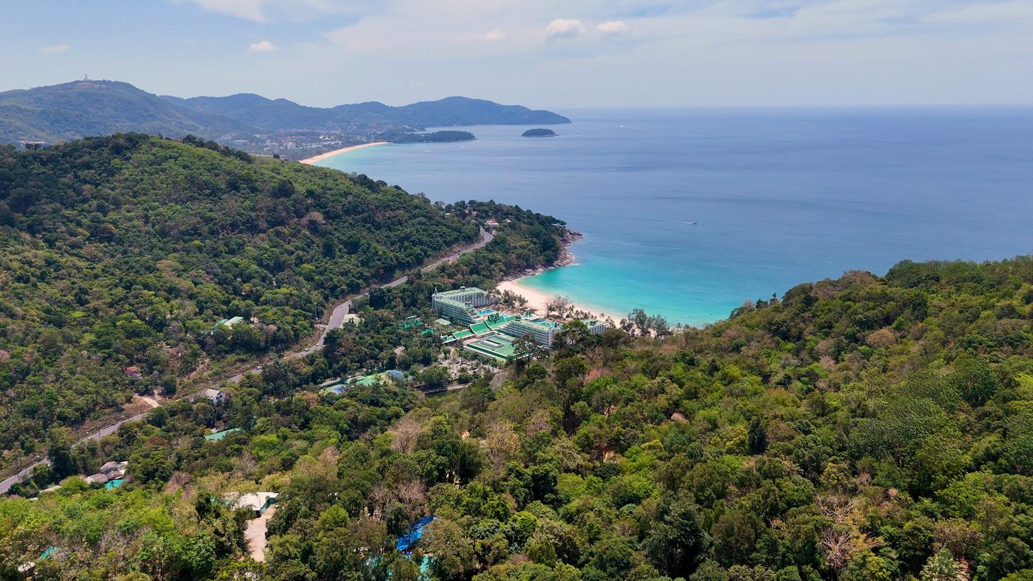 An aerial view of low-rise resort buildings on a wooded hillside above a Phuket bay