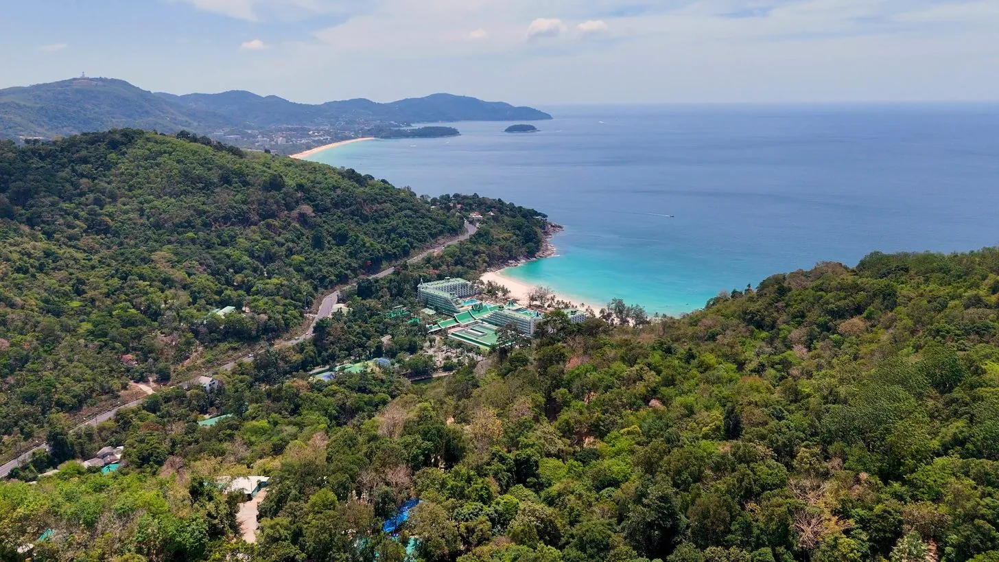 Luftaufnahme niedriger Resortgebäude an einem bewaldeten Hang über einer Bucht auf Phuket