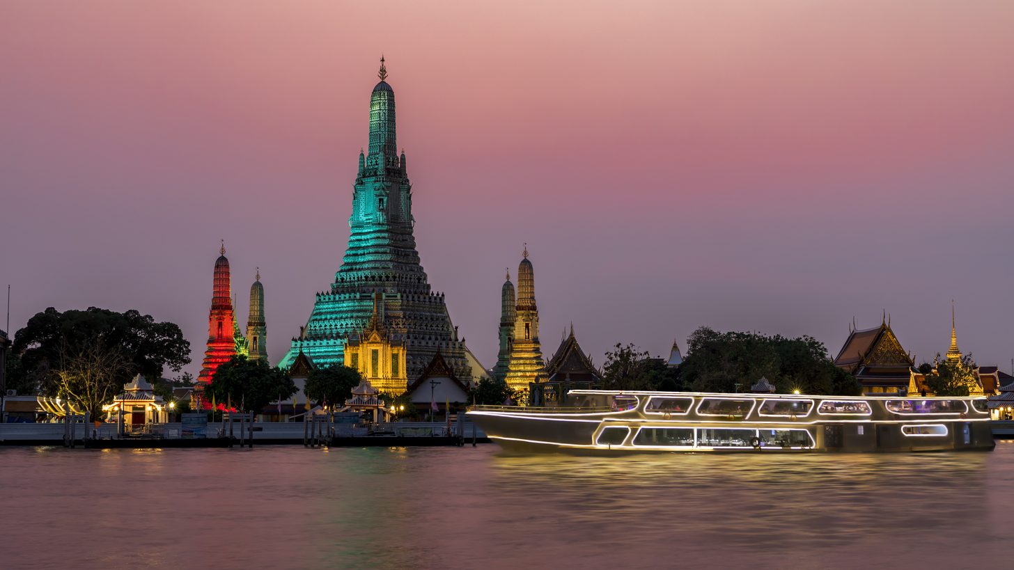Wat Arun lit at dusk above the Chao Phraya with an illuminated river boat passing