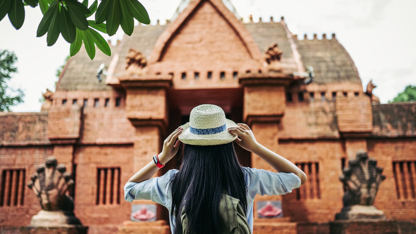 A visitor standing before the entrance of a laterite temple, hands raised to her hat