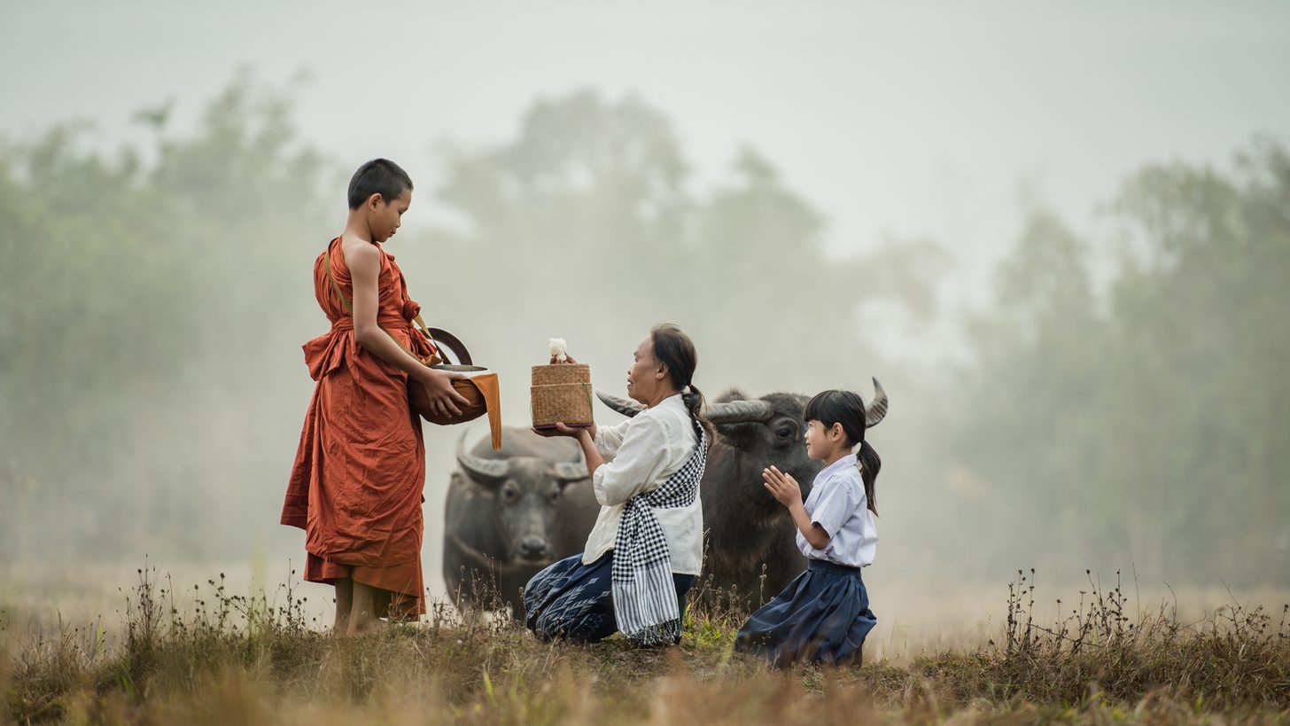 An older woman kneeling to offer food to a young Buddhist novice in a misty field, a child beside her with palms pressed together