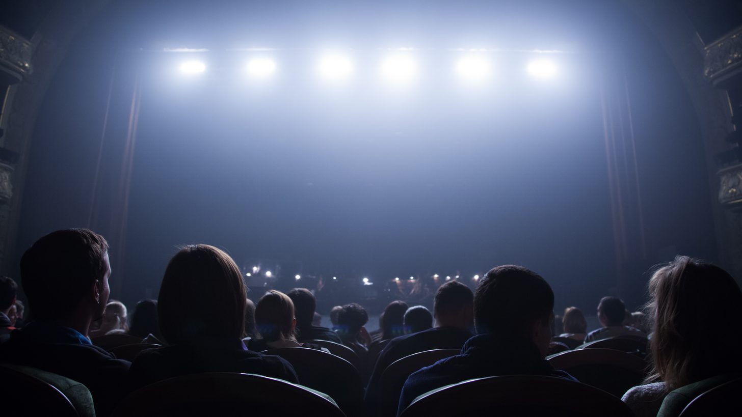 An audience seated in a darkened theatre facing a brightly lit stage