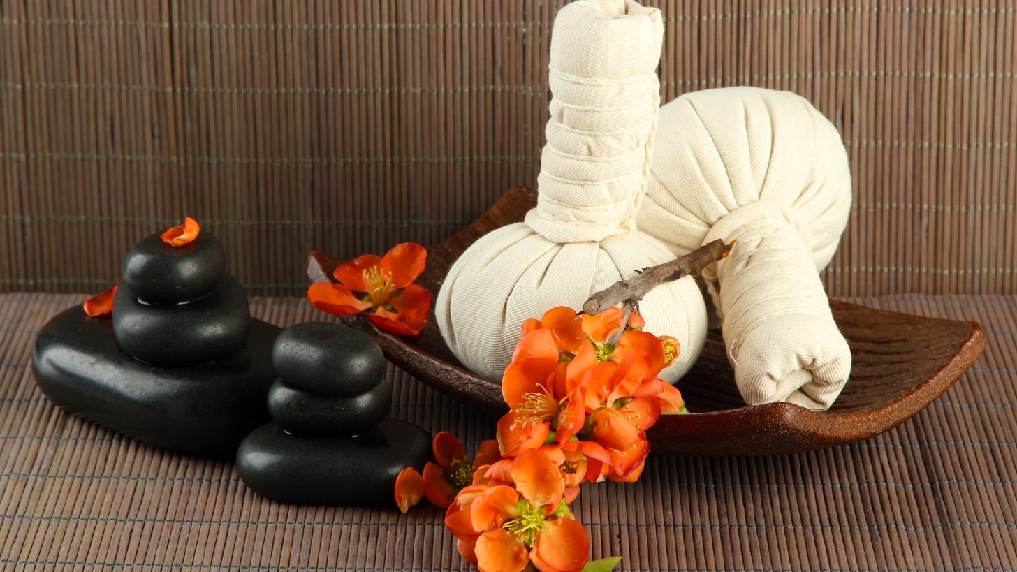 Herbal compress balls on a wooden tray with massage stones on a bamboo mat