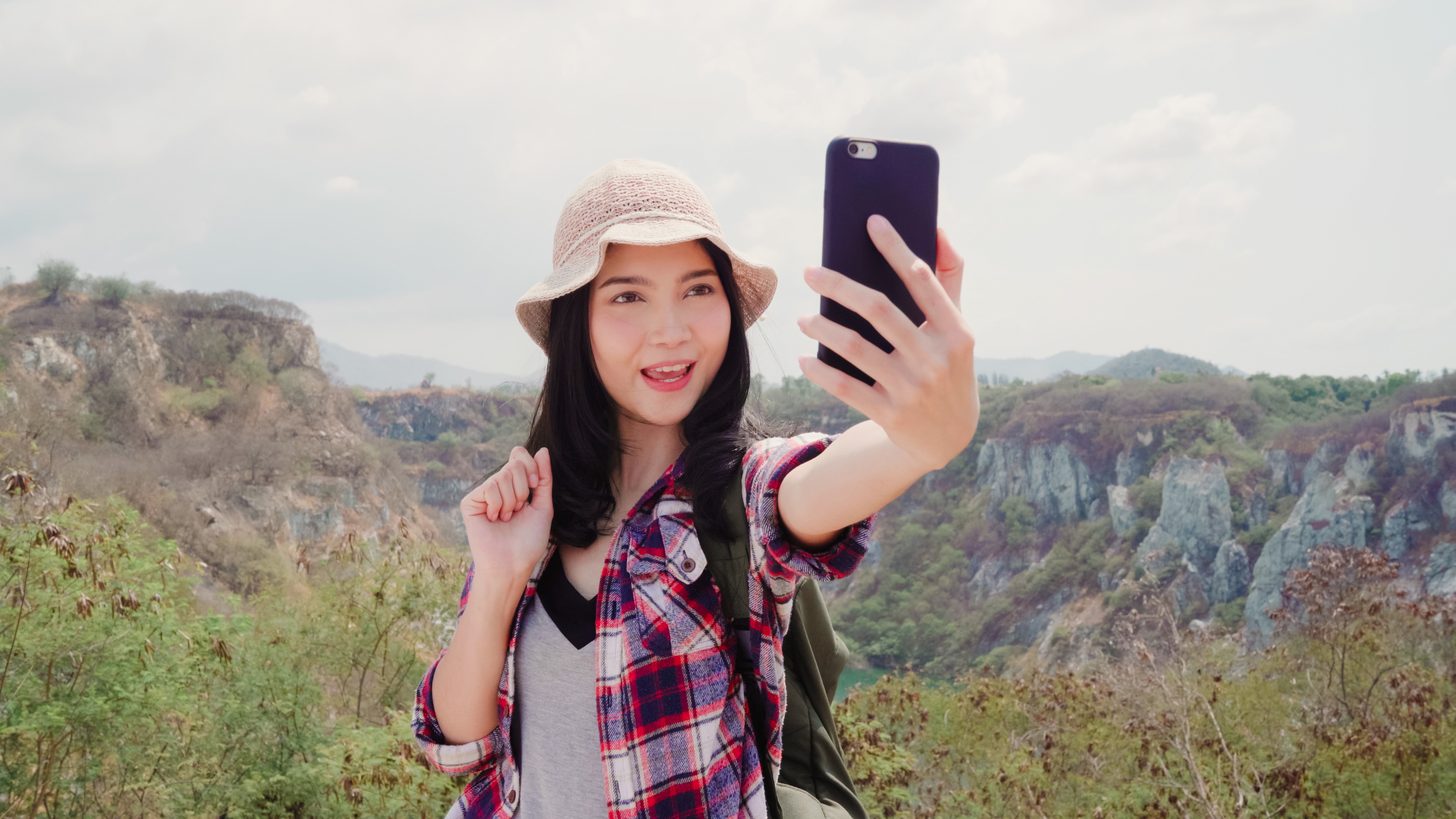 A traveller in a sun hat filming herself on a phone at a hilltop viewpoint