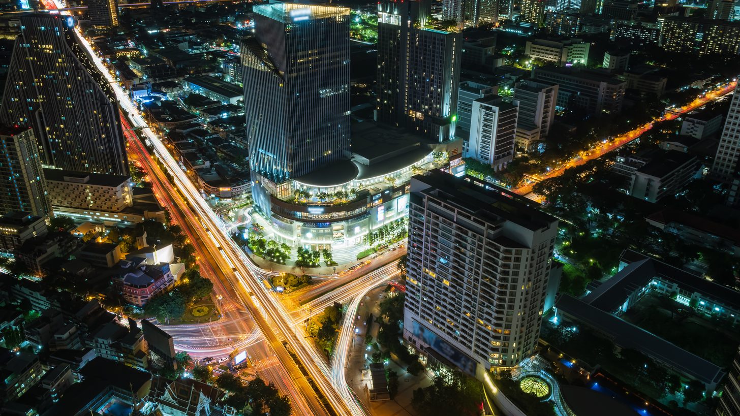 Aerial night view of a Bangkok business district intersection with towers, hotels and traffic trails