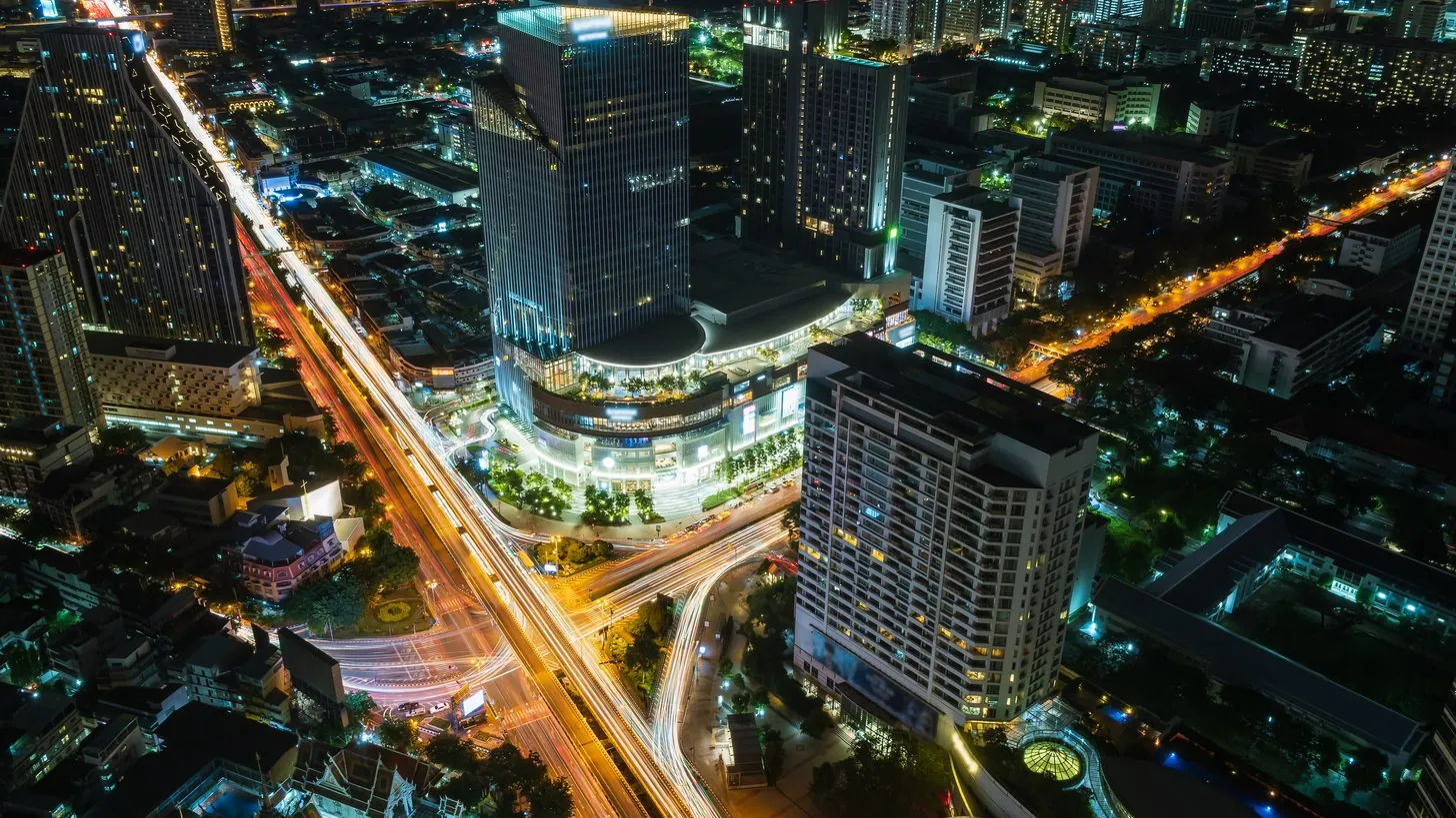 Nächtliche Luftaufnahme einer Kreuzung im Bangkoker Geschäftsviertel mit Türmen, Hotels und Lichtspuren des Verkehrs