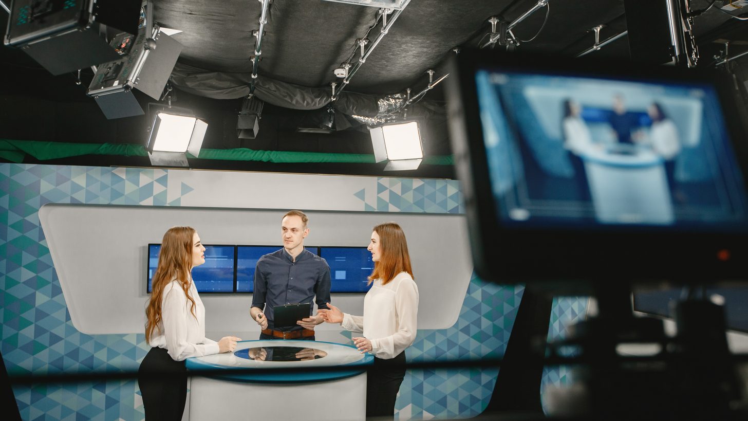 Two presenters and a guest around a desk on a lit television studio set, a camera monitor in the foreground