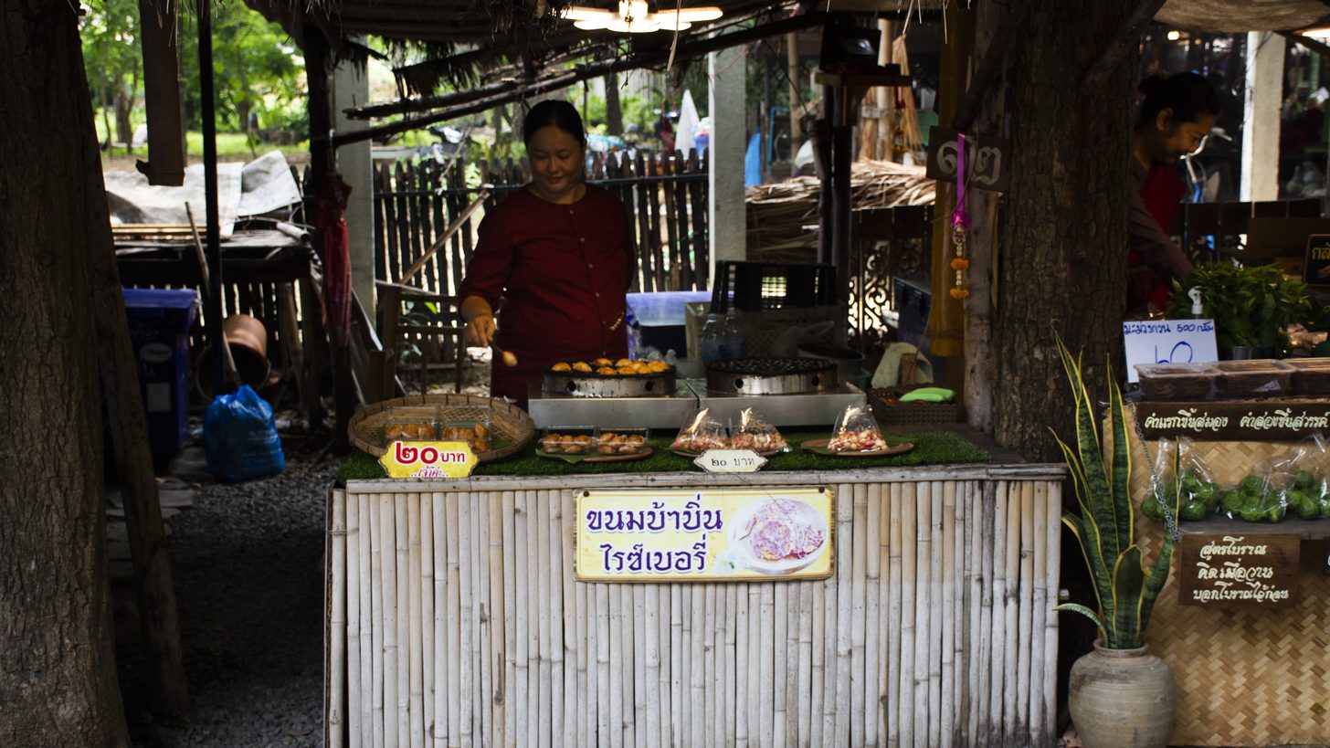 A woman cooking a single traditional Thai sweet at a bamboo roadside stall, handwritten Thai signs propped along the counter
