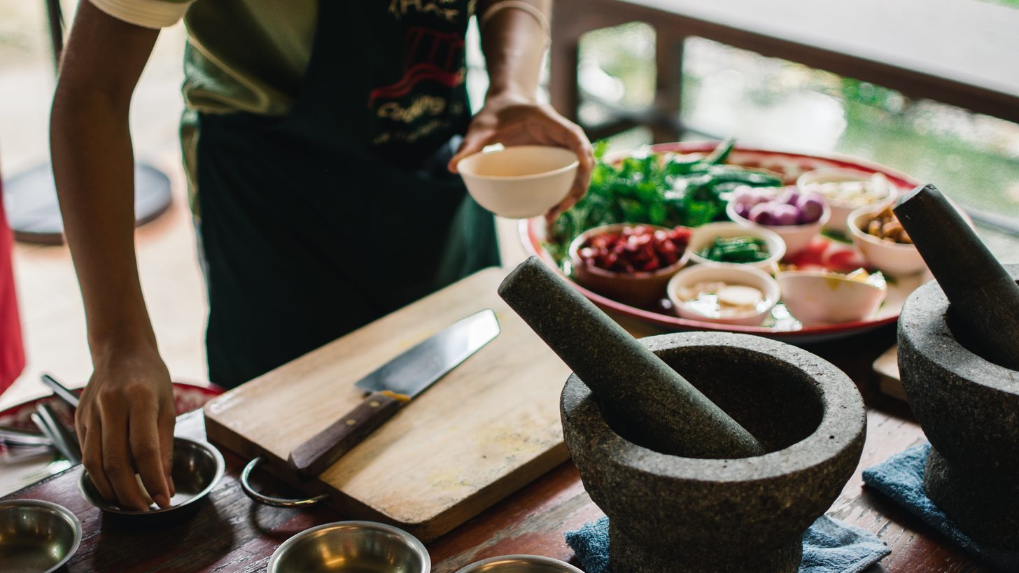 A Thai cooking class in progress: stone mortars and pestles, a Thai cleaver on a board, and prepped ingredients in small bowls on a tray