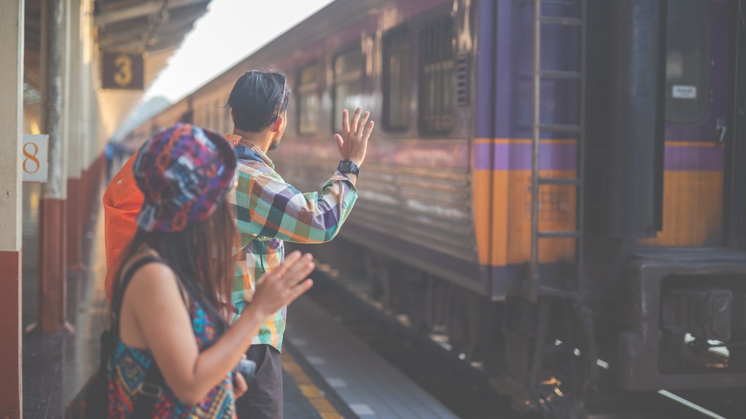 Two travellers with backpacks waving from a Thai station platform as a State Railway carriage pulls away