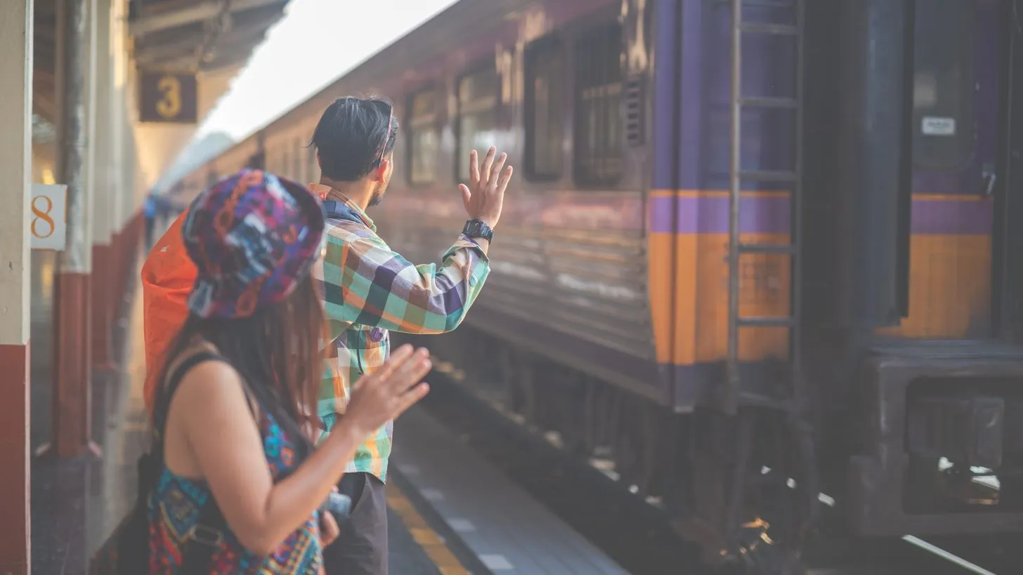 Zwei Reisende mit Rucksäcken winken von einem thailändischen Bahnsteig, während ein Wagen der State Railway abfährt