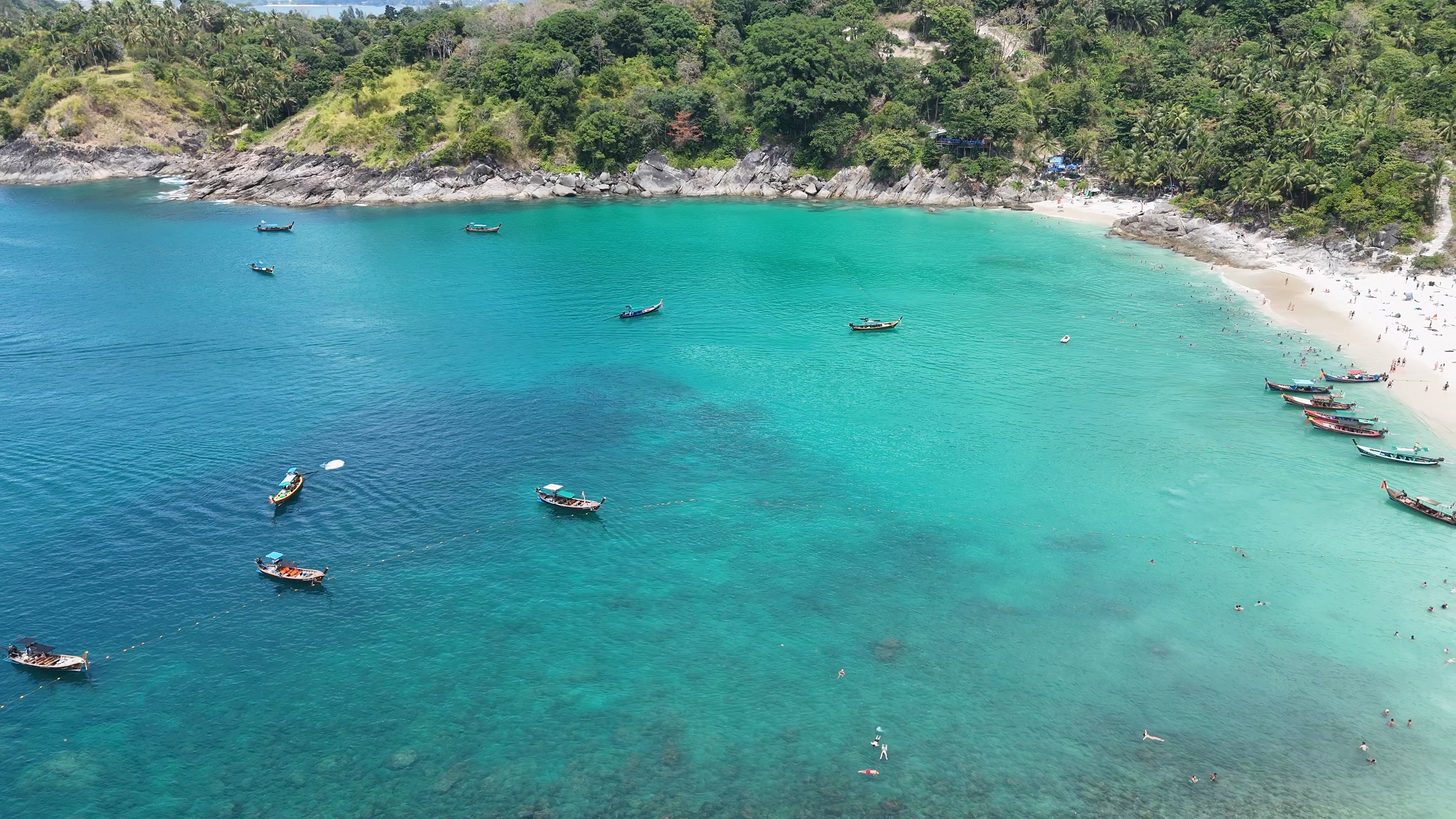 Aerial view of a Phuket bay with longtail boats moored over turquoise shallows and a forested headland behind the beach