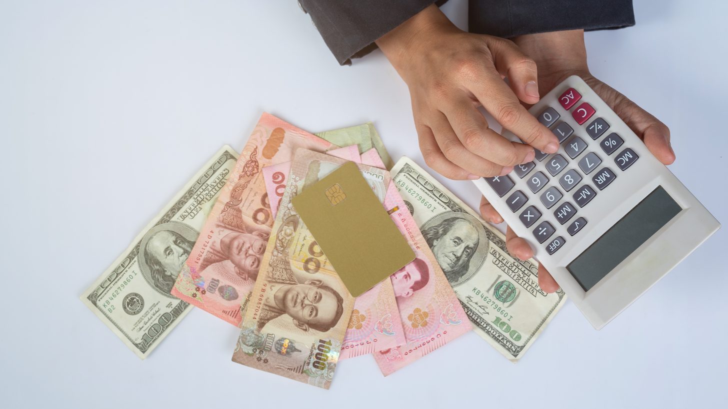 Thai baht and US dollar notes fanned out beside a payment card and a calculator, with a hand entering a figure