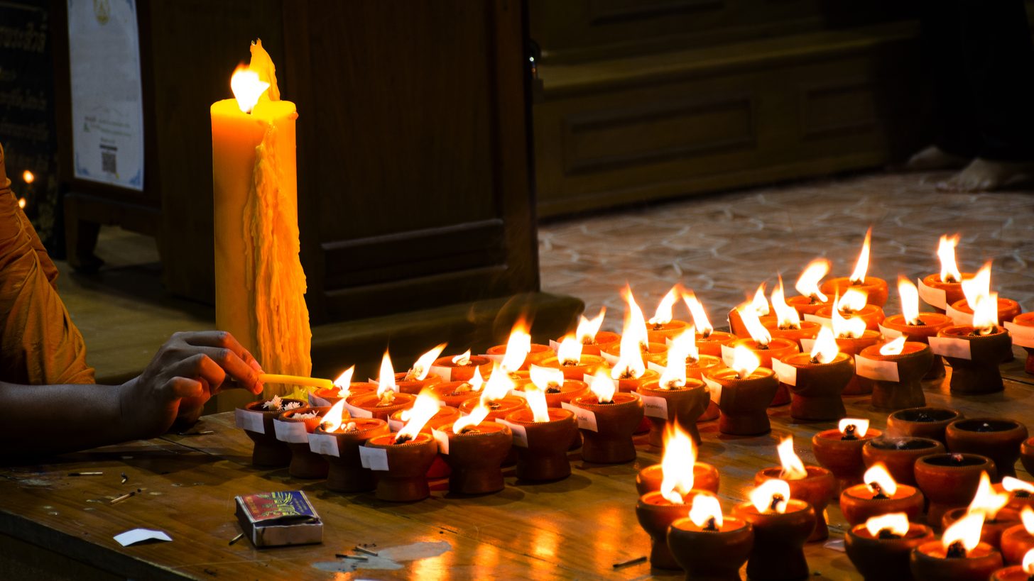 Ein Mönch in safranfarbener Robe entzündet abends Reihen von Öllampen in einem thailändischen Tempel