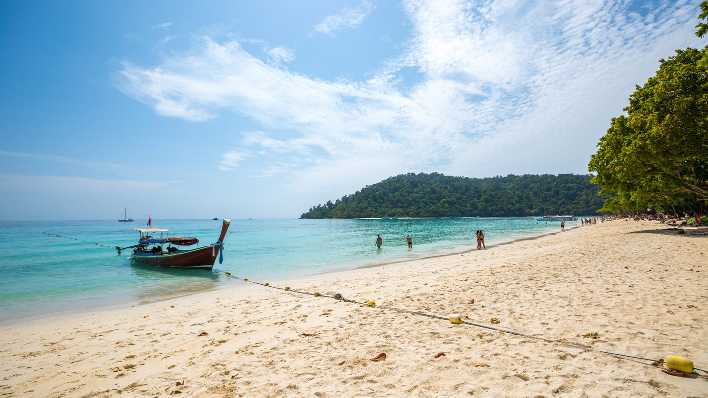 A wooden longtail boat moored on a white-sand Krabi beach with a forested headland across the bay