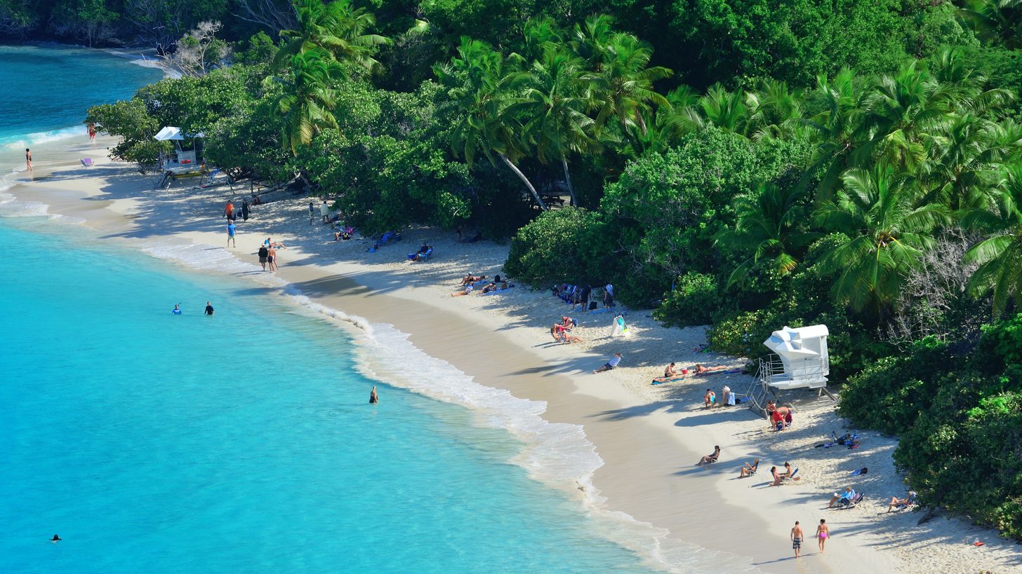 A wide tropical beach of pale sand meeting clear turquoise water, with green headlands behind