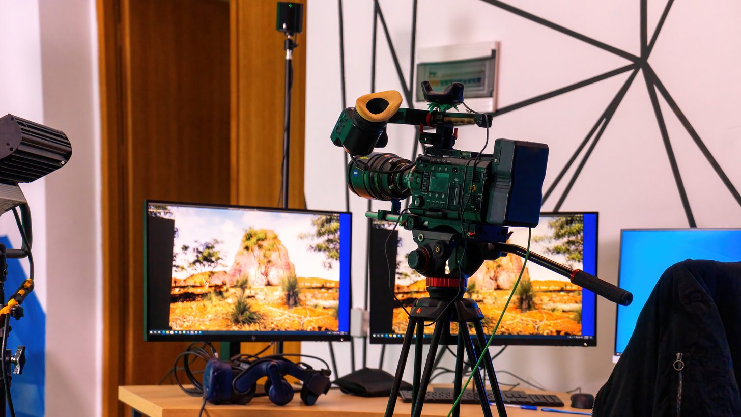A professional video camera on a stand beside monitors in a studio