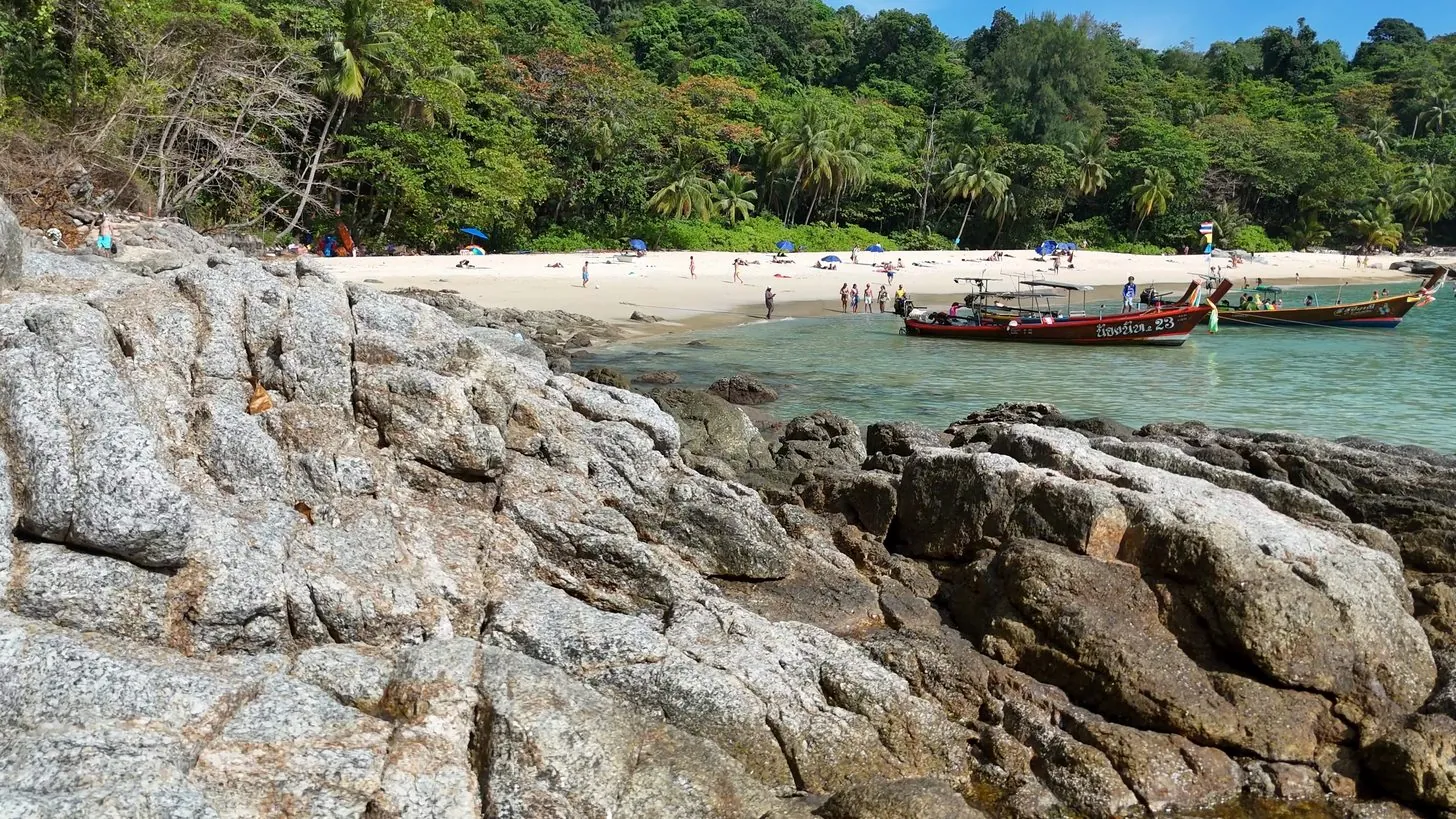 Longtail-Boote vor einem Strand auf Phuket, hinter einem felsigen Ufer