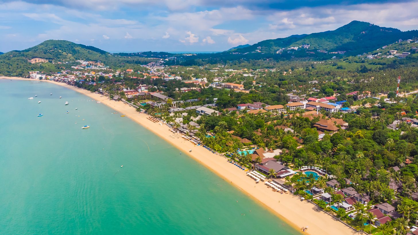 Vista aérea de uma longa praia de areia em Koh Samui com resorts entre palmeiras e colinas verdes ao fundo