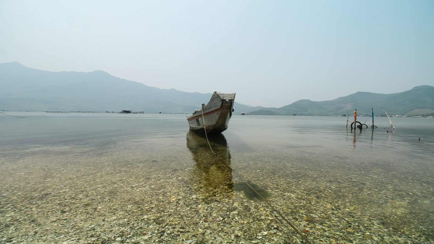 Ein hölzernes Fischerboot liegt im flachen Wasser an der Küste Zentralvietnams vor einer Hügelkette
