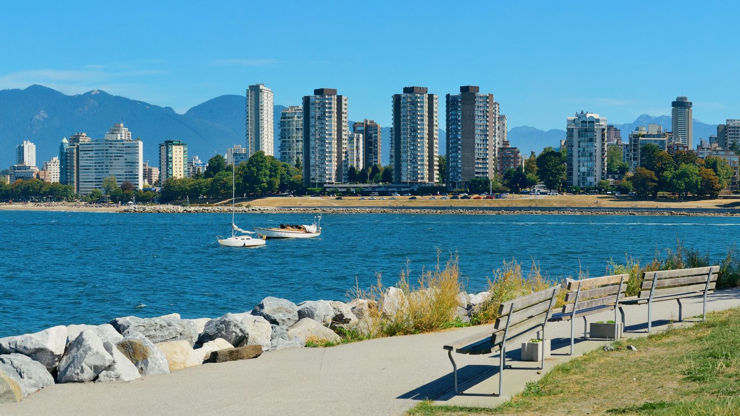 Downtown Vancouver's apartment towers across the water, with mountains behind and benches on the waterfront