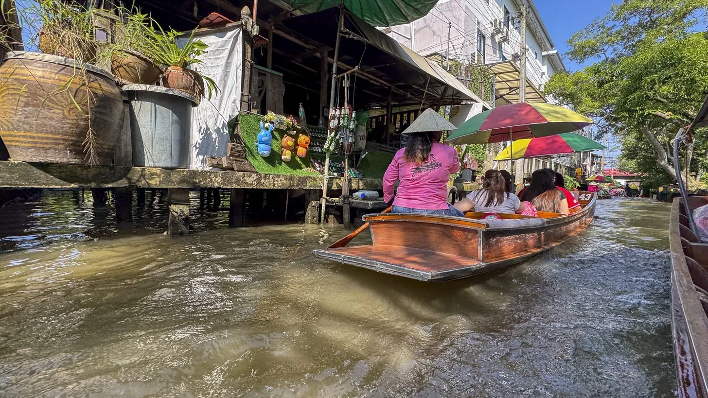 Holzboote mit Besuchern auf einem von Häusern am Wasser gesäumten Kanal nahe Bangkok
