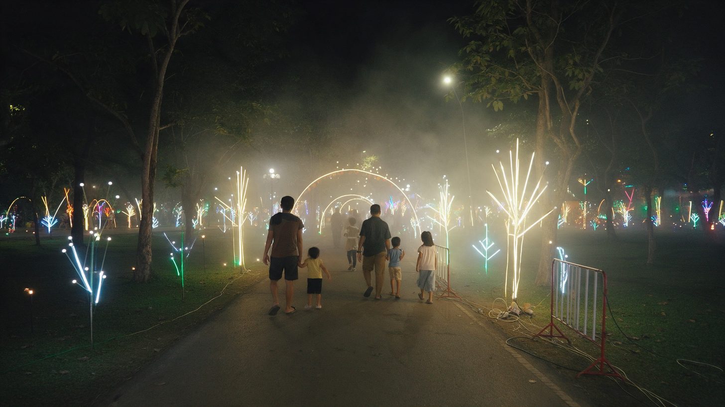 ครอบครัวเดินบนทางเดินในสวนสาธารณะที่ประดับงานแสงไฟเรียบง่ายในคืนที่อากาศอบอ้าว