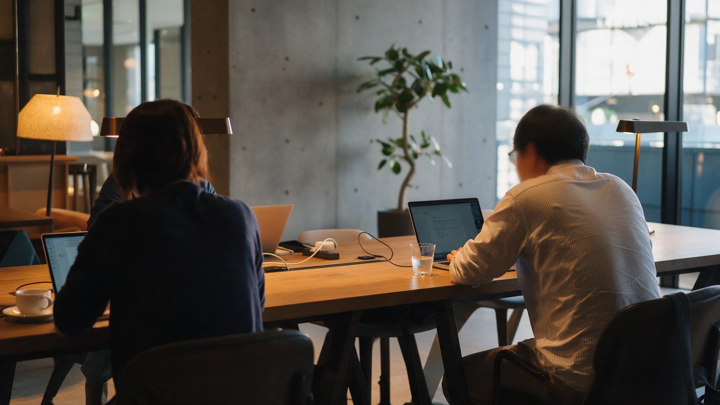 Zwei Personen arbeiten an Laptops an einem langen gemeinsamen Tisch in einer Hotellounge