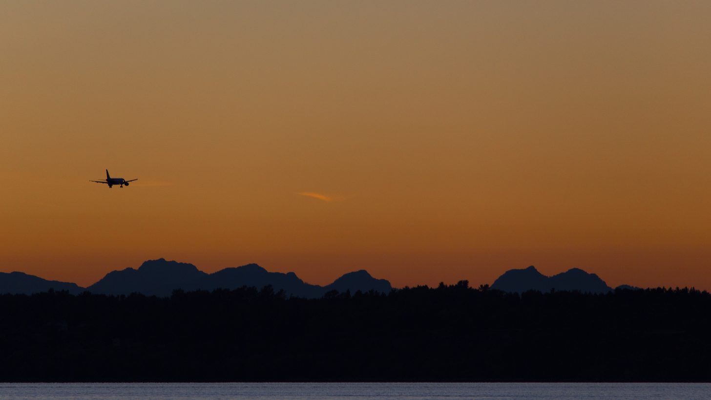 An aircraft silhouetted against an orange evening sky above a dark ridge of hills