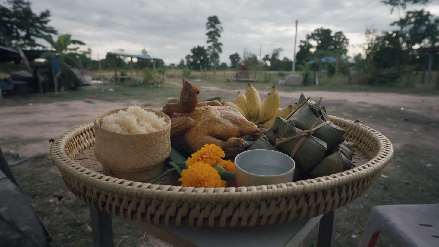 A woven bamboo tray of offerings with sticky rice, a roasted chicken, bananas and marigolds on a table outdoors