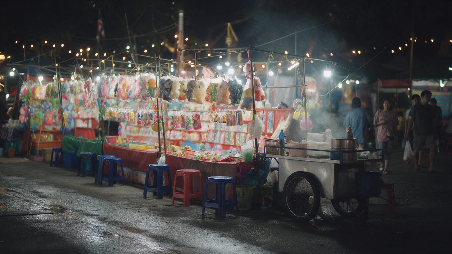 Game stalls with hanging prizes under string lights at a night fair, with plastic stools and a food cart in front