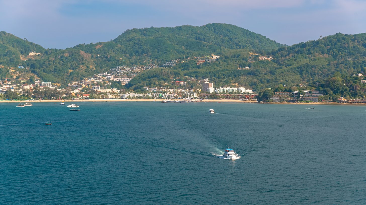 Boats sailing past a coastal town on Phuket, with green hills behind