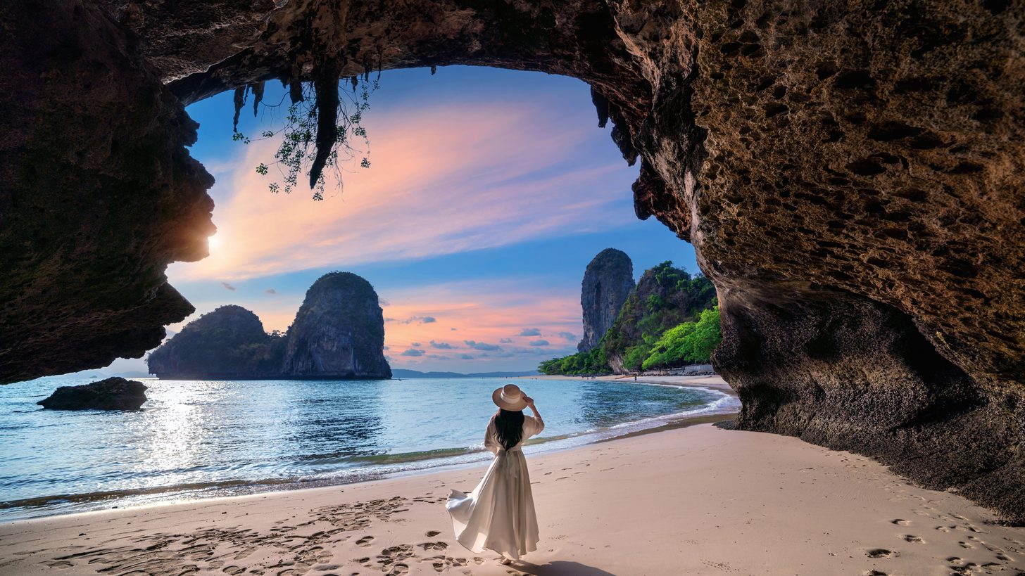 A woman walking on Railay beach in Krabi, framed by a cave and limestone stacks