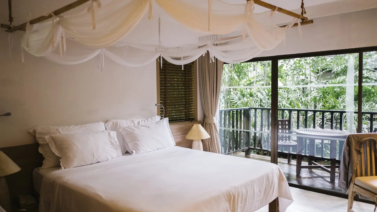 Ein Hotelzimmer mit weißem Bett unter einem drapierten Baldachin und einem Balkon mit Blick auf Bäume