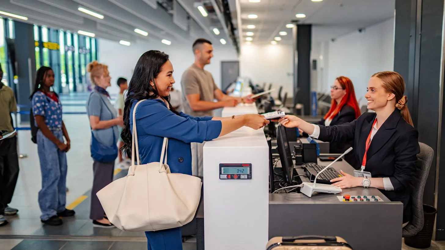 A traveller hands her passport to an agent at an airline check-in counter, other passengers queuing behind